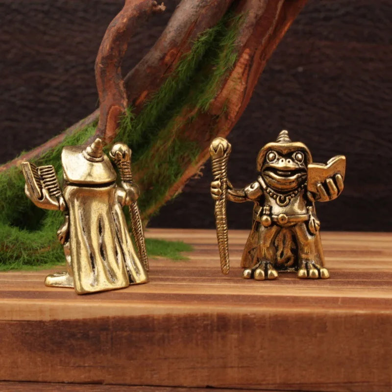 Two gold frog figurines of warrior-like creatures on a wooden surface with a natural background.