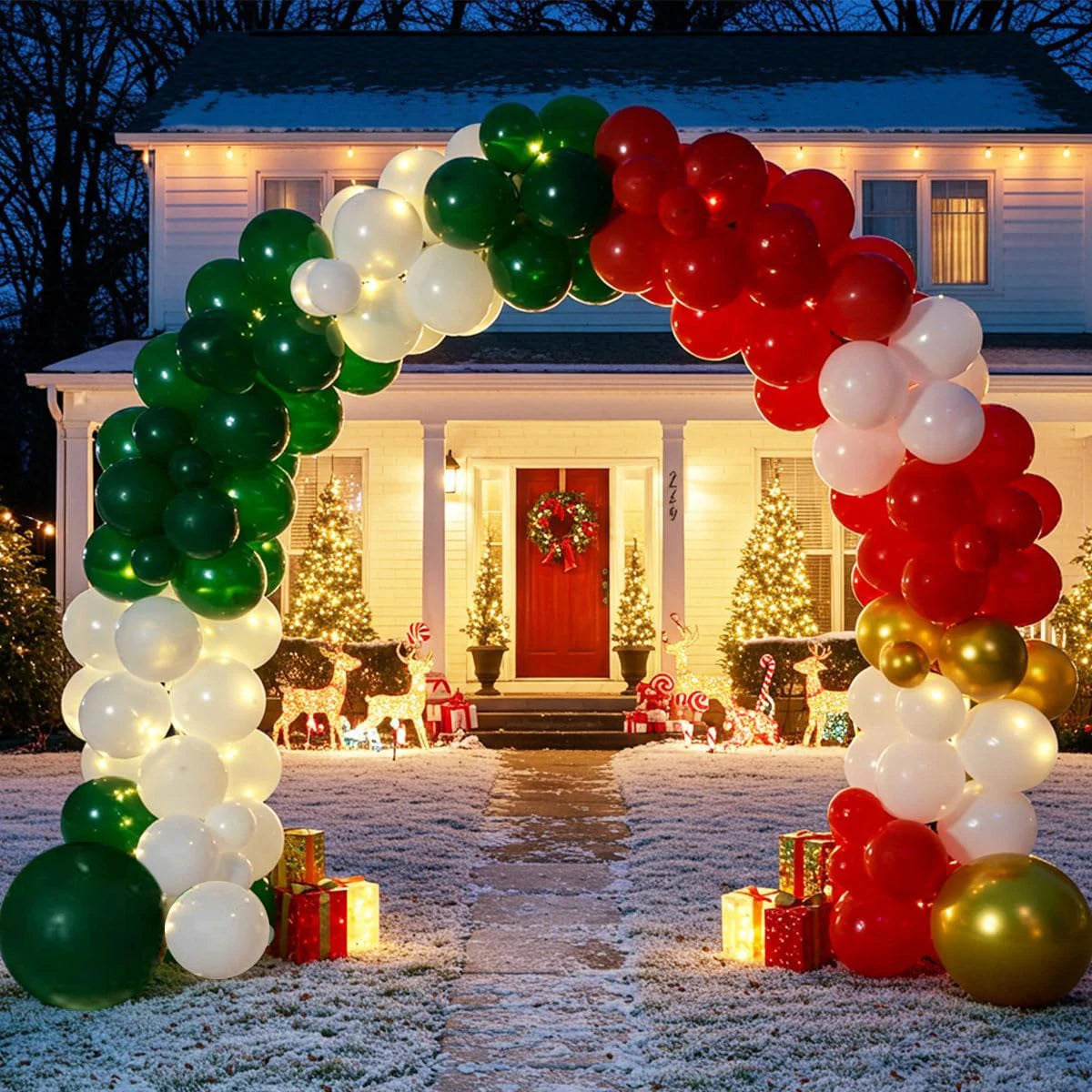 A festive holiday balloon arch made of green, white, red, and gold balloons framing the front porch of a house at night. The porch is decorated with Christmas trees, lights, gifts, and a wreath, with snow lightly dusting the ground.