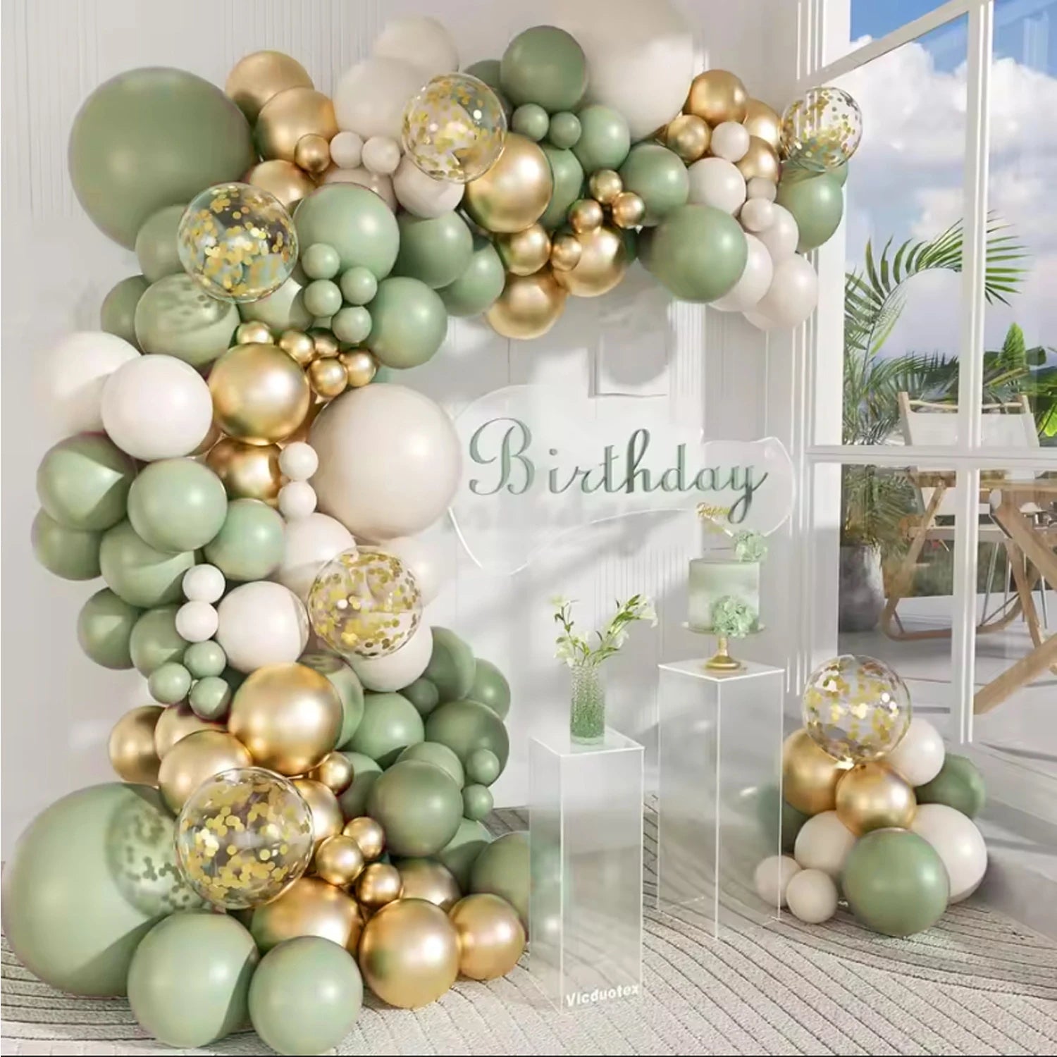 A large balloon garland arch decorated in sage green, metallic gold, and cream, with clear balloons filled with gold confetti. The setup is styled as a birthday party backdrop with a 'Birthday' sign, clear acrylic tables, and a window showing a bright outdoor patio scene.