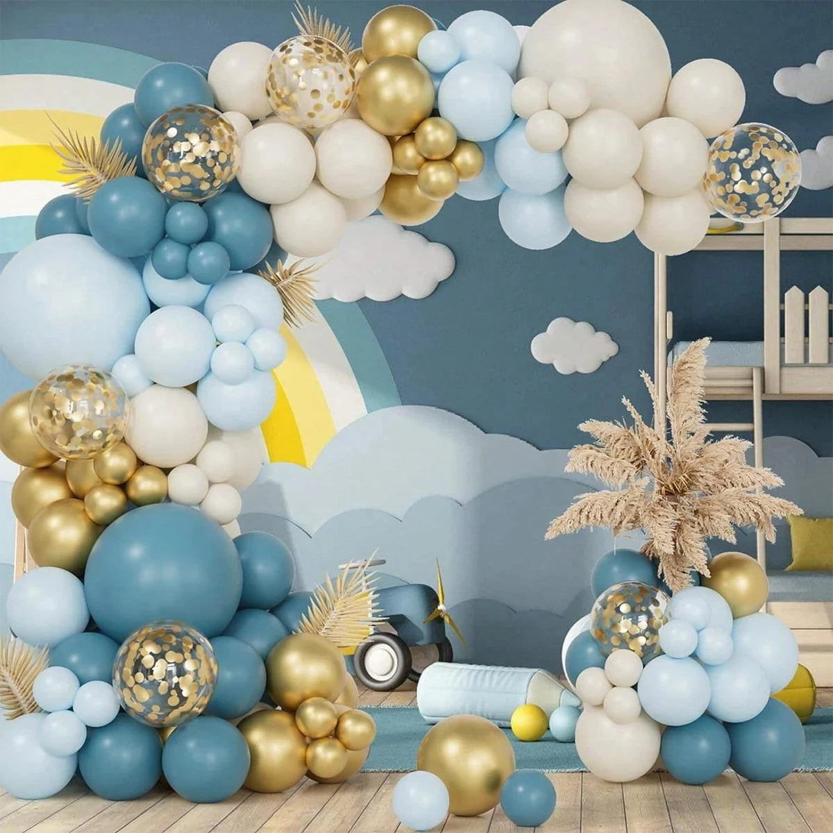 A decorative balloon garland arch and floor clusters in pastel blue, cream, white, and metallic gold, styled in a children's room with cloud decals, a small toy car, and dried grass decor.