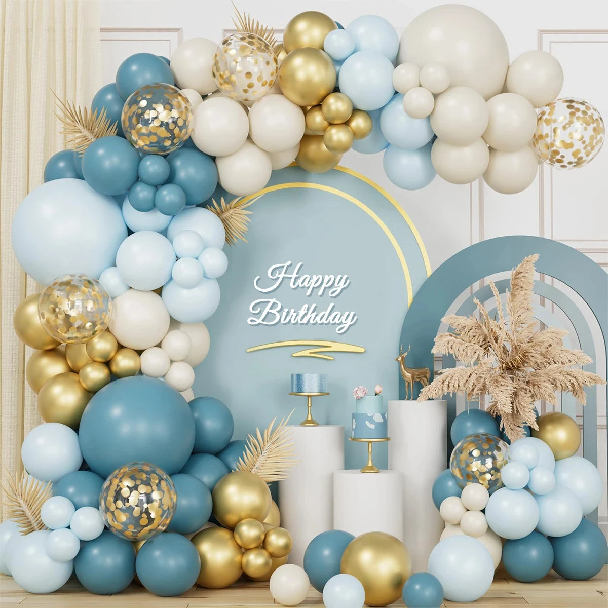 A festive birthday party backdrop featuring a large balloon garland arch in light blue, cream, and metallic gold, with a central blue arch and 'Happy Birthday' sign, plus small cake stands and decorative dried grass.