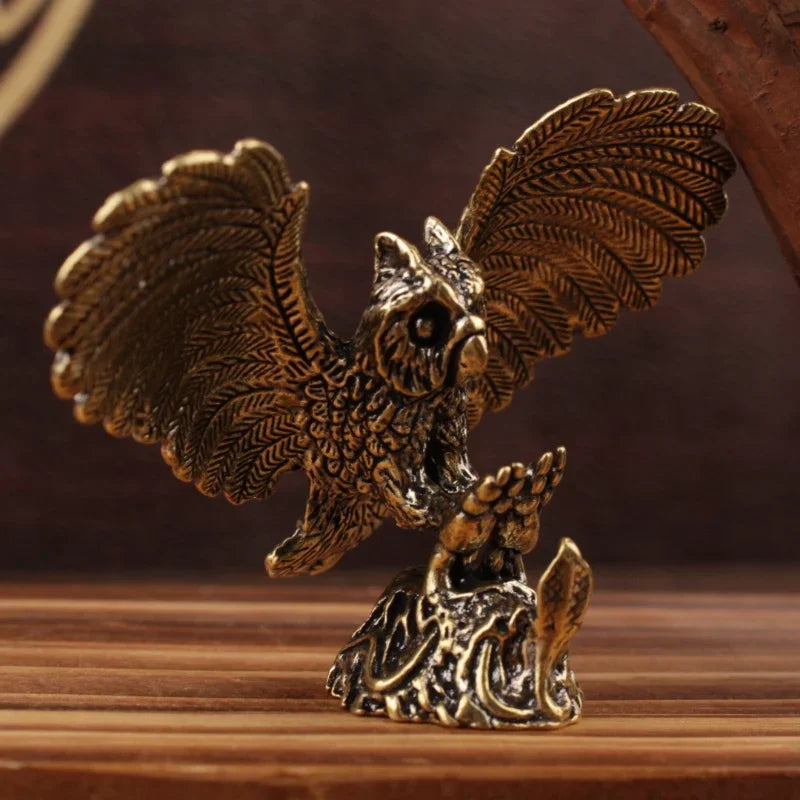 A bronze-finished metal owl figurine with wings spread, perched on a textured base, displayed on a wooden surface against a dark brown background.