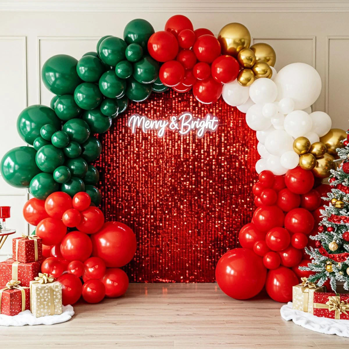 Christmas-themed balloon garland backdrop with green, red, white, and gold balloons framing a red sequin wall with 'Merry & Bright' text, decorated with gift boxes and a small Christmas tree.