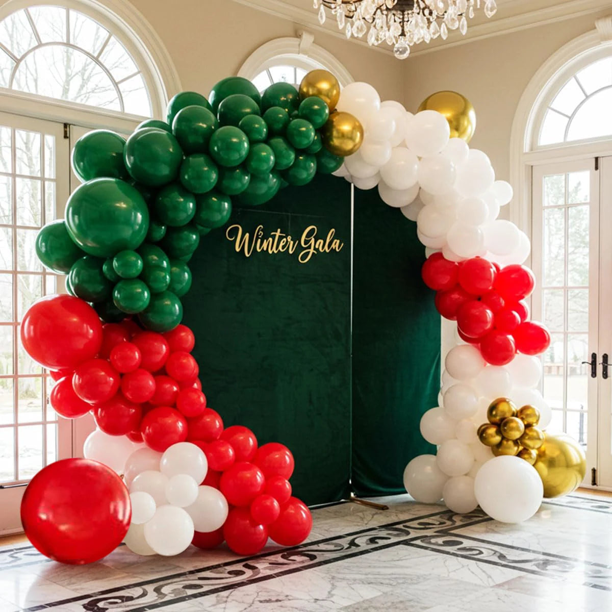 A festive balloon arch backdrop for a 'Winter Gala' event, featuring clusters of green, red, and white balloons with gold accents, set against a dark green velvet curtain in a grand indoor hall with arched windows.