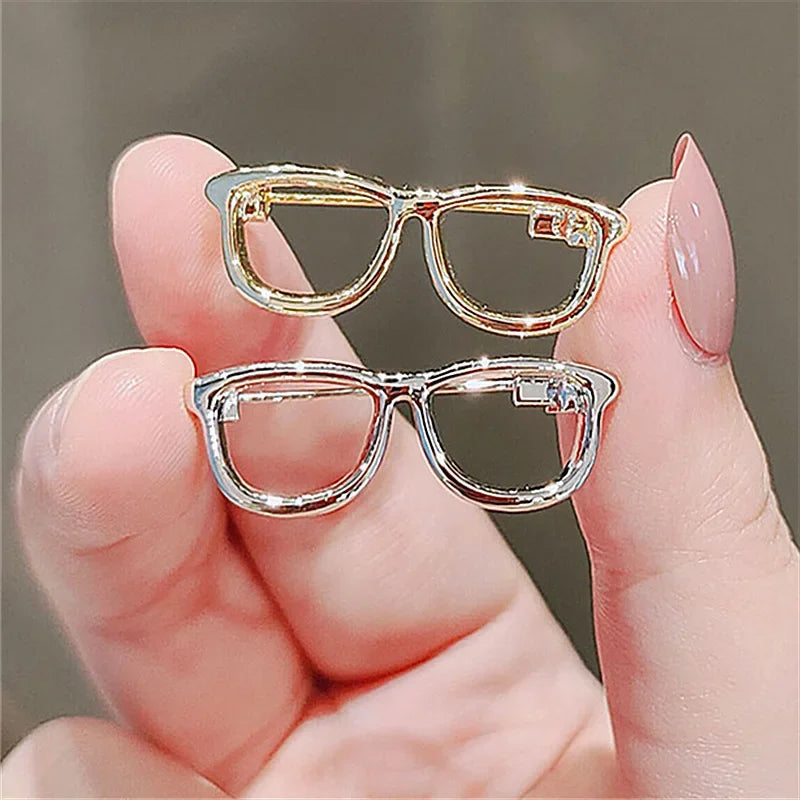Gold and silver glasses brooch pins held between fingers on neutral background