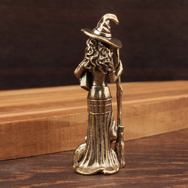A small bronze-finished witch figurine standing on a dark wooden surface with a wooden block in the background.