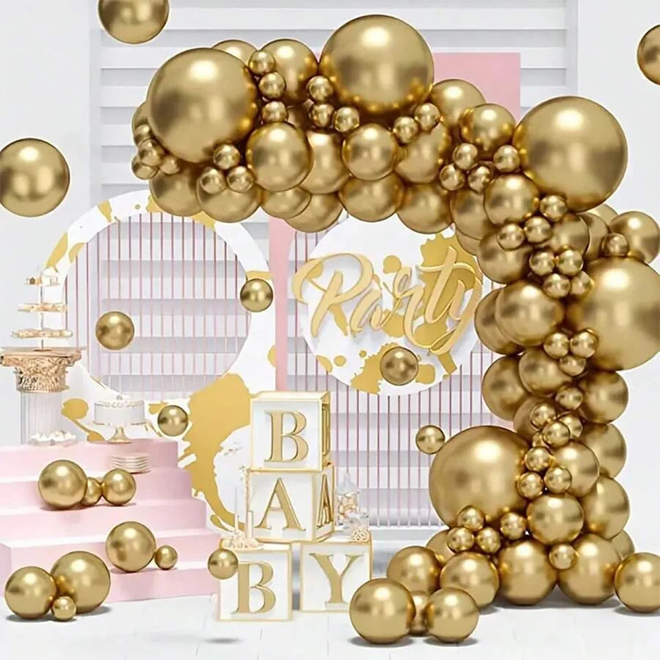 Gold balloon garland arch decoration with metallic gold balloons in various sizes, arranged in a party setup with pink and white striped backdrop, gold 'Party' script sign, stacked 'BABY' blocks, and a small cake stand.