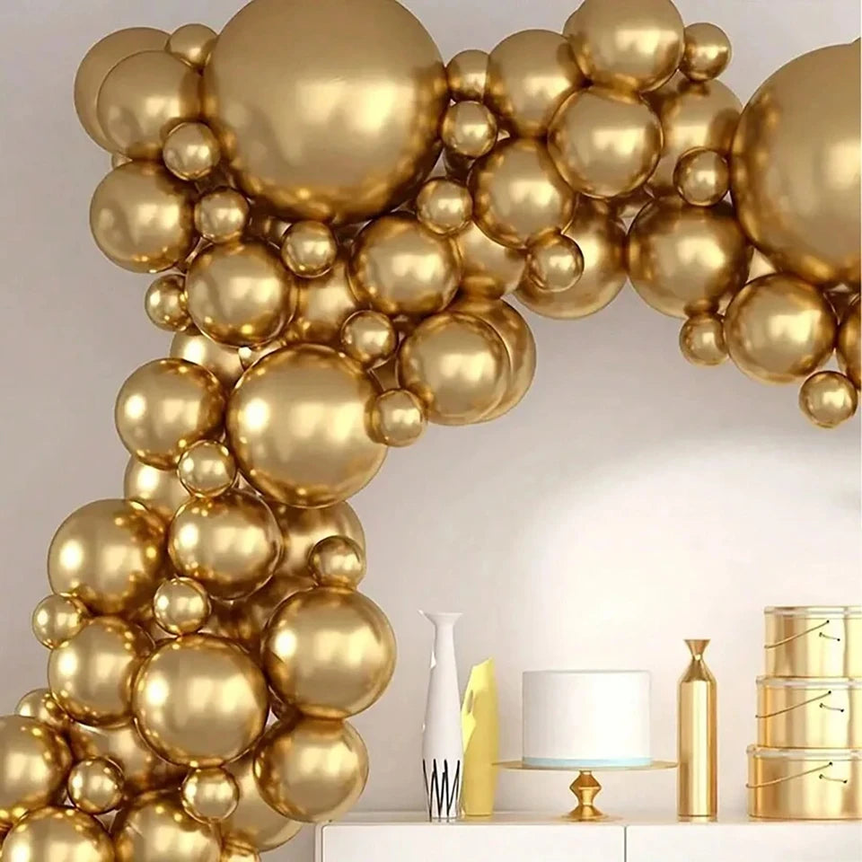 A decorative balloon garland made of many metallic gold balloons in assorted sizes, arranged in a curved arch against a light neutral wall. A white console table with decorative objects is visible below.