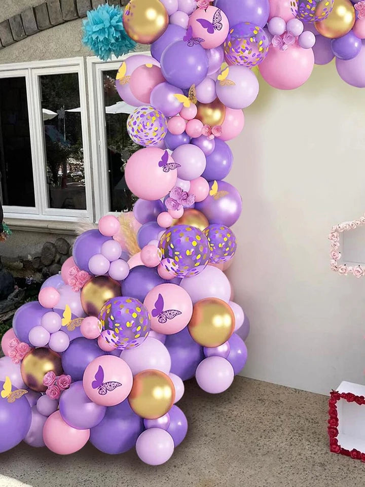 A decorative balloon garland arrangement in pastel pink, lavender, and metallic gold, featuring butterfly and floral accents, set up against an outdoor patio wall.