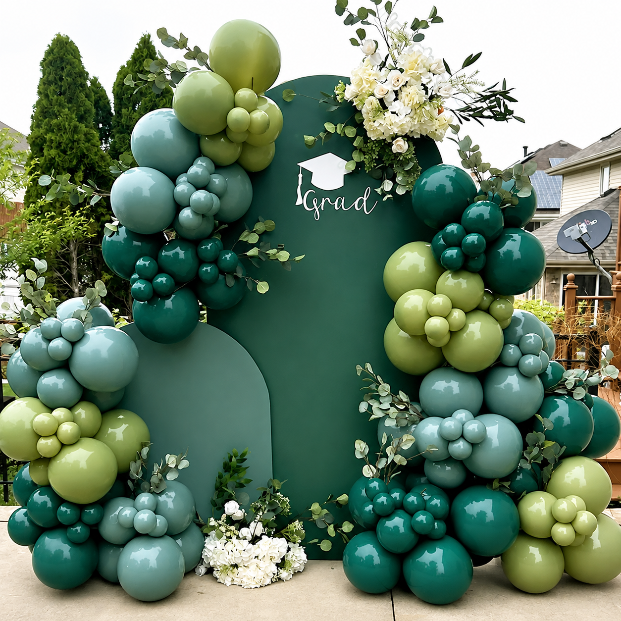 A graduation party backdrop setup featuring a large dark green arch with clusters of glossy balloons in teal, mint green, and sage green, accented with white and green flowers. The word 'grad' is printed in white script on the center of the backdrop, with a small graduation cap icon above it.