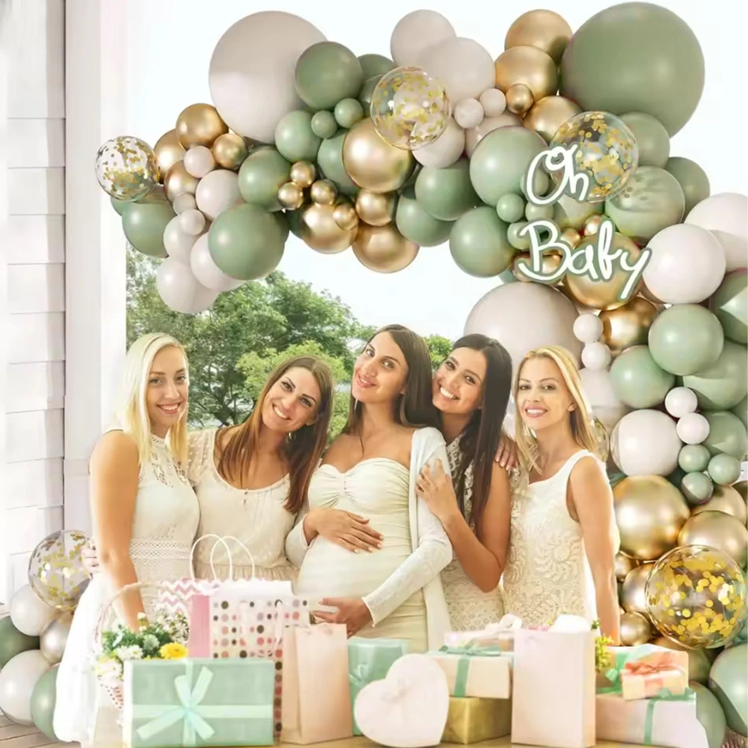 A group of five pregnant women posing together in front of a large balloon garland arch decorated with sage green, white, and gold balloons, along with gold and silver confetti balloons. The women are dressed in white maternity outfits, and the scene is decorated with baby gift bags and wrapped presents on a table in the foreground.