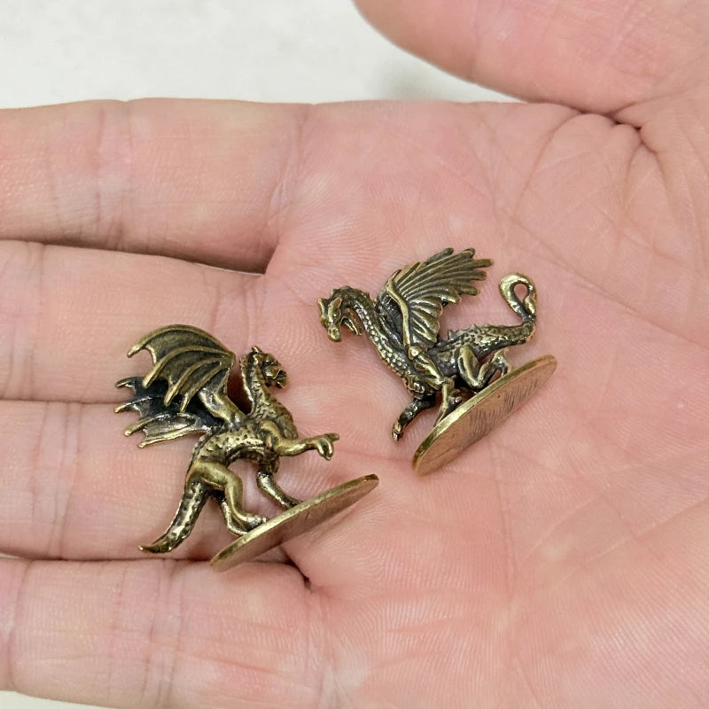 Two small winged dragon figurines in an antique gold finish, displayed on a person's palm to show scale.