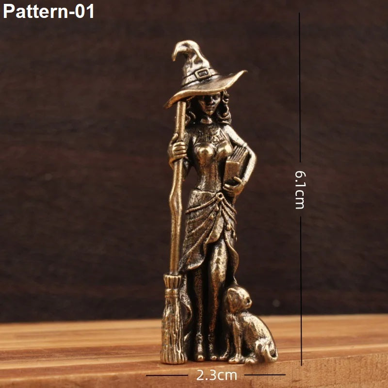 A small bronze-colored witch figurine holding a broom and book, shown on a wooden surface against a dark brown background, with dimension labels indicating 6.1 cm height and 2.