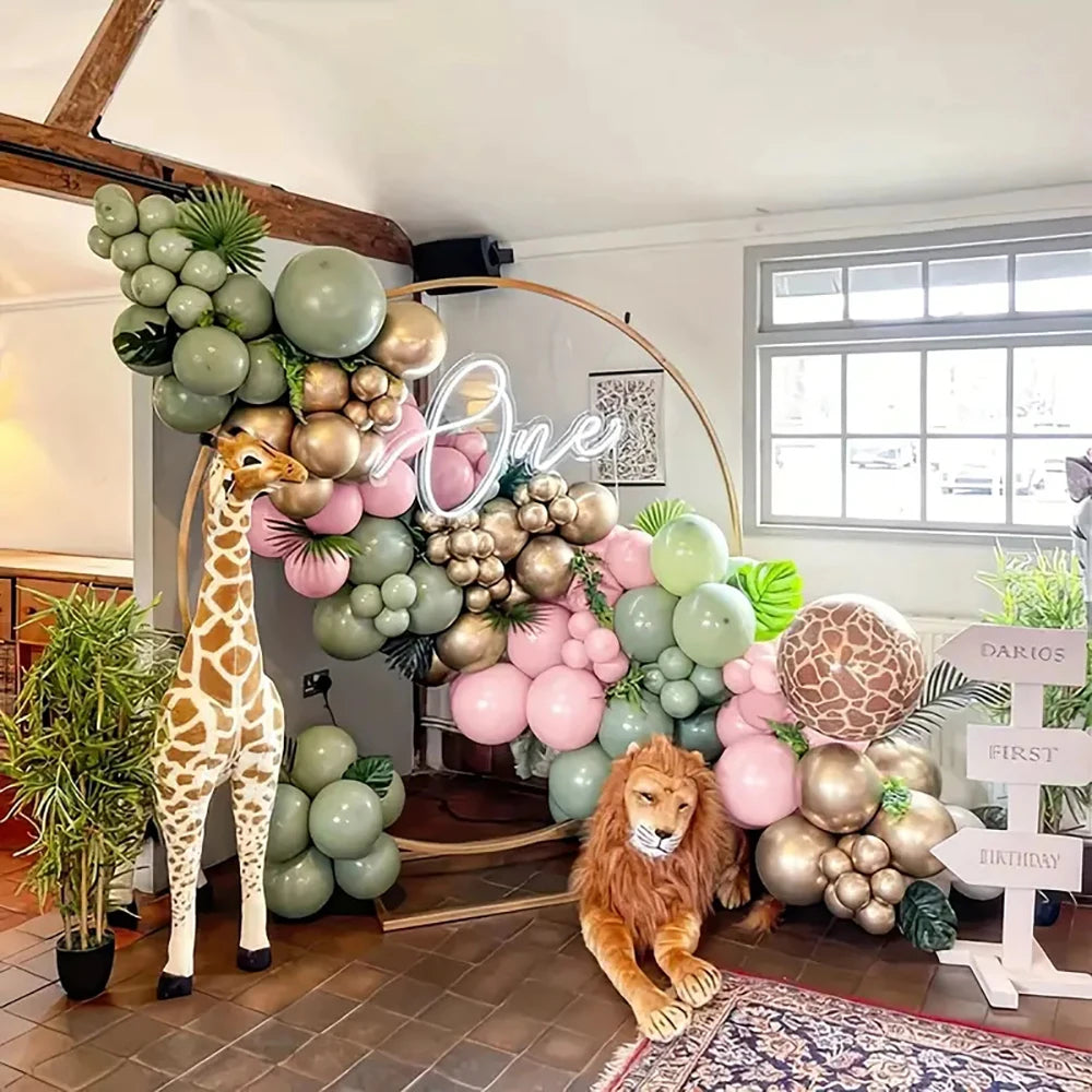 A decorated first birthday party setup in a room with a large balloon arch in sage green, blush pink, and metallic gold, a giraffe decoration, a lion plush, and a white sign reading 'DARIO'S FIRST BIRTHDAY JUNE 2026'.