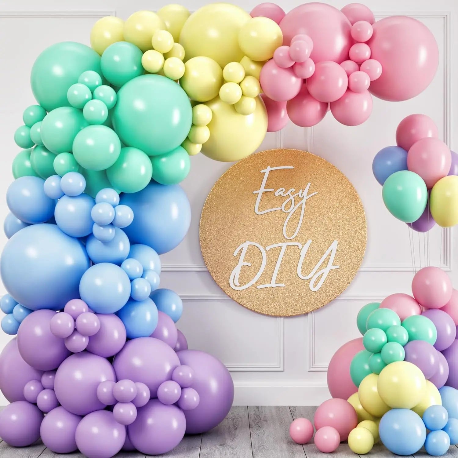 A pastel rainbow balloon garland arch displayed on a white paneled wall, with a round gold sign reading 'Easy DIY' in the center. The balloons come in pink, yellow, mint green, light blue, and lavender purple, with some extra balloons clustered on the floor and to the side.