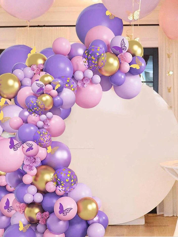 Purple, pink, and gold balloon garland arch decoration with butterfly and floral accents, set against a white circular backdrop in an indoor event space.