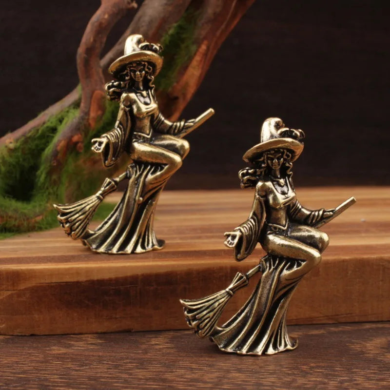 Two bronze witch figurines on brooms on a wooden surface with a natural background.