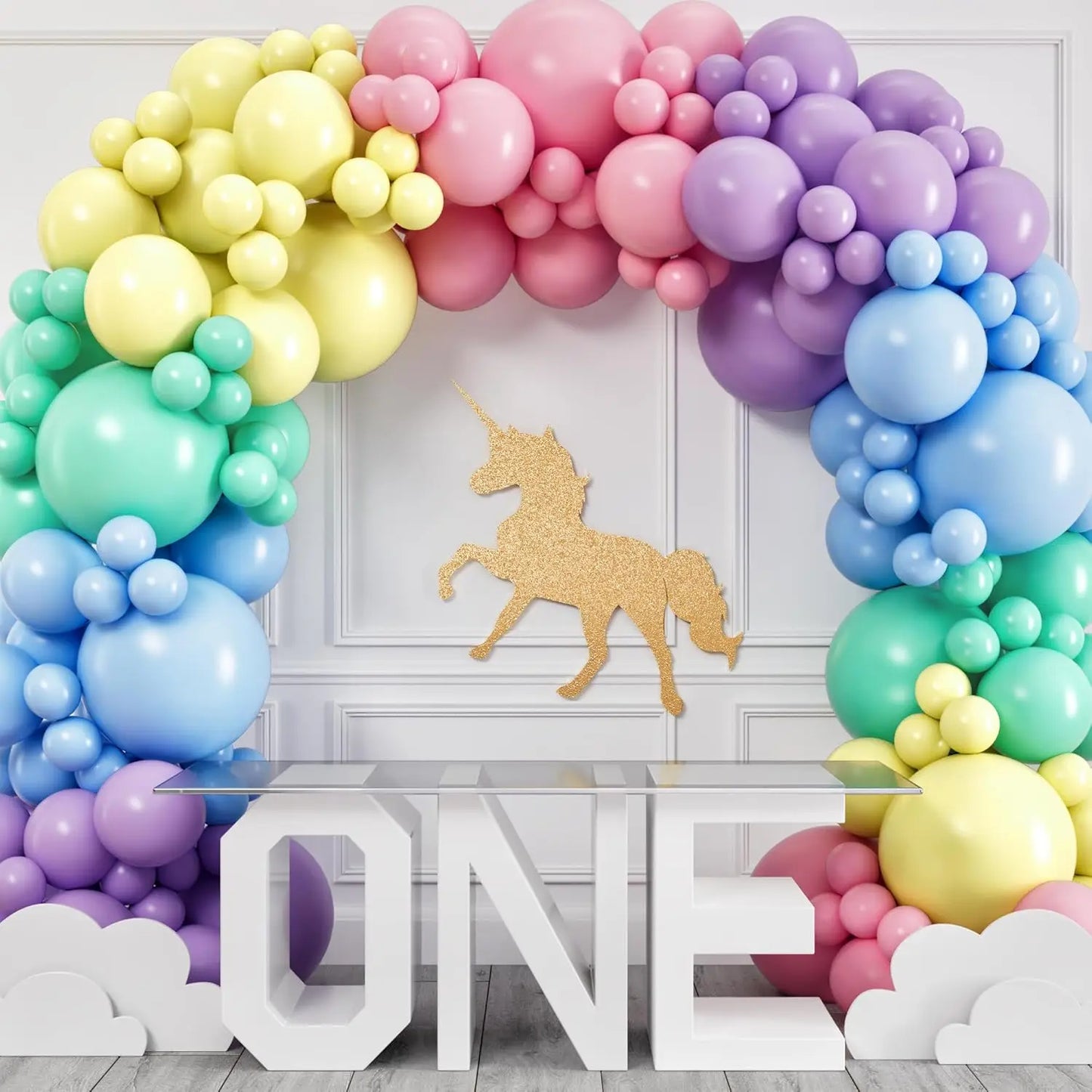 Colorful balloon arch with a gold unicorn cutout and 'ONE' letter on a white wall.