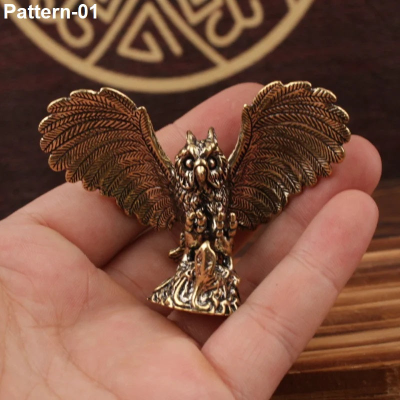A bronze-colored metal owl figurine with spread wings, held in a person's hand. The background shows a dark wooden surface with a circular gold-toned design and the text 'Pattern-01' in the top left corner.