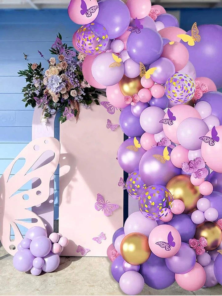 A decorative party backdrop featuring a large balloon garland in pastel pink, lavender, and metallic gold, with butterfly decorations, a butterfly-shaped panel, and floral accents against a blue paneled background.