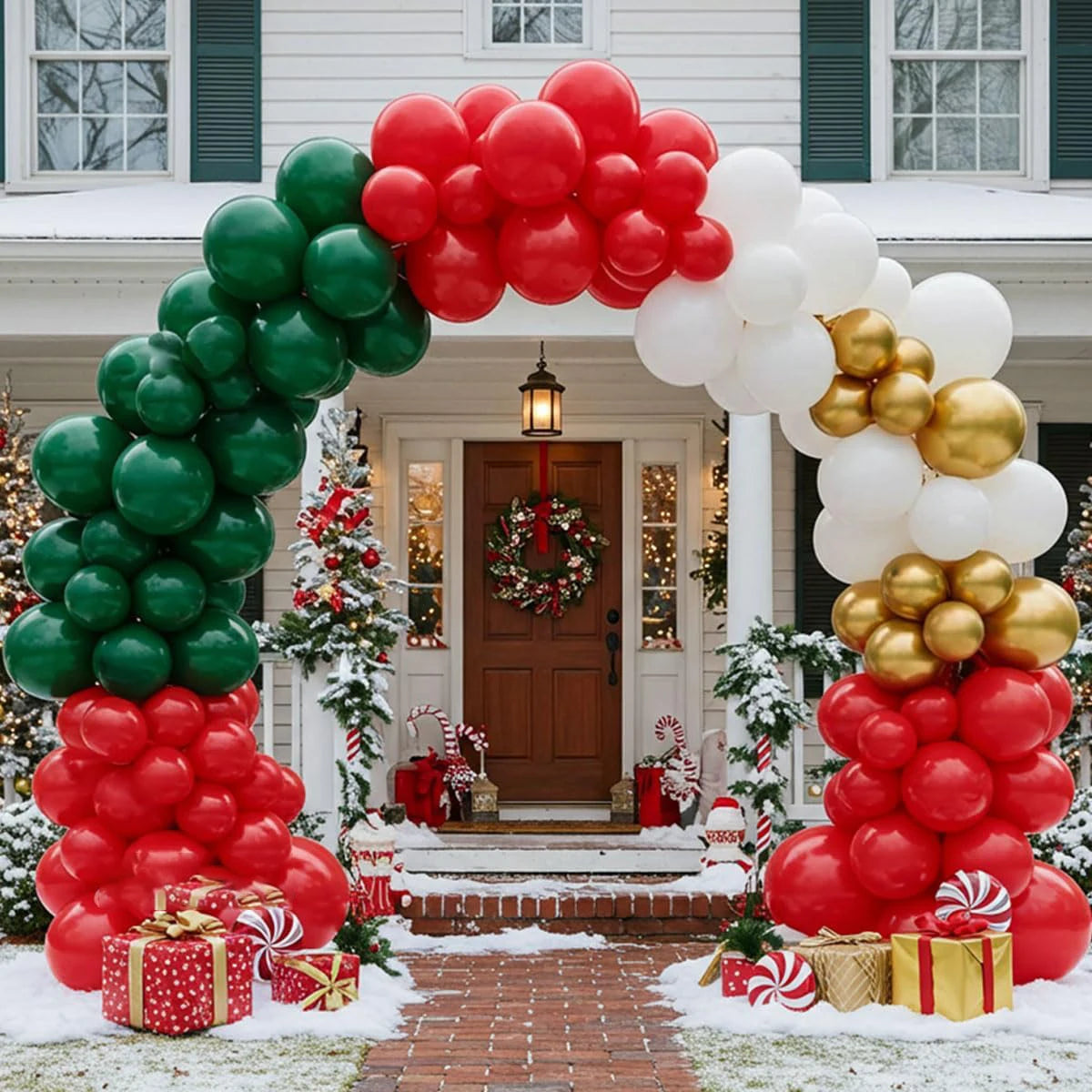A festive Christmas balloon arch decoration set up in front of a house. The arch features large balloons in red, green, white, and metallic gold, with matching balloon clusters resting on the ground and wrapped gift boxes nearby.