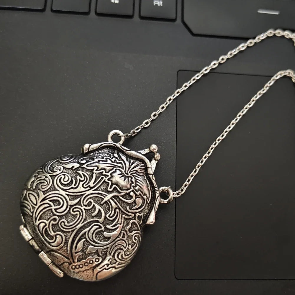 An ornate silver locket pendant necklace with intricate floral engravings, resting on a black laptop keyboard.