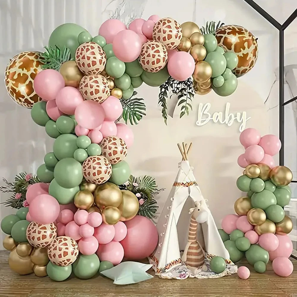 A baby shower party decoration setup featuring a large balloon arch and matching side clusters in pastel pink, sage green, metallic gold, and giraffe-print patterns, with a small teepee and a baby sign in the center.