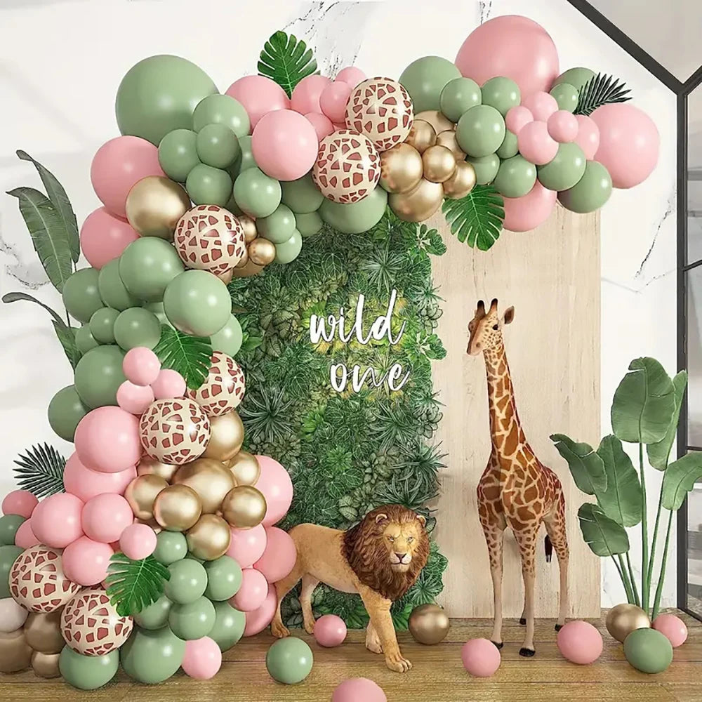 A jungle-themed party decoration setup featuring a large balloon arch in pastel pink, sage green, and metallic gold, with patterned balloons, faux greenery, a faux wood backdrop, and toy animals including a giraffe and lion. The words 'wild one' are written in white script on the greenery backdrop.