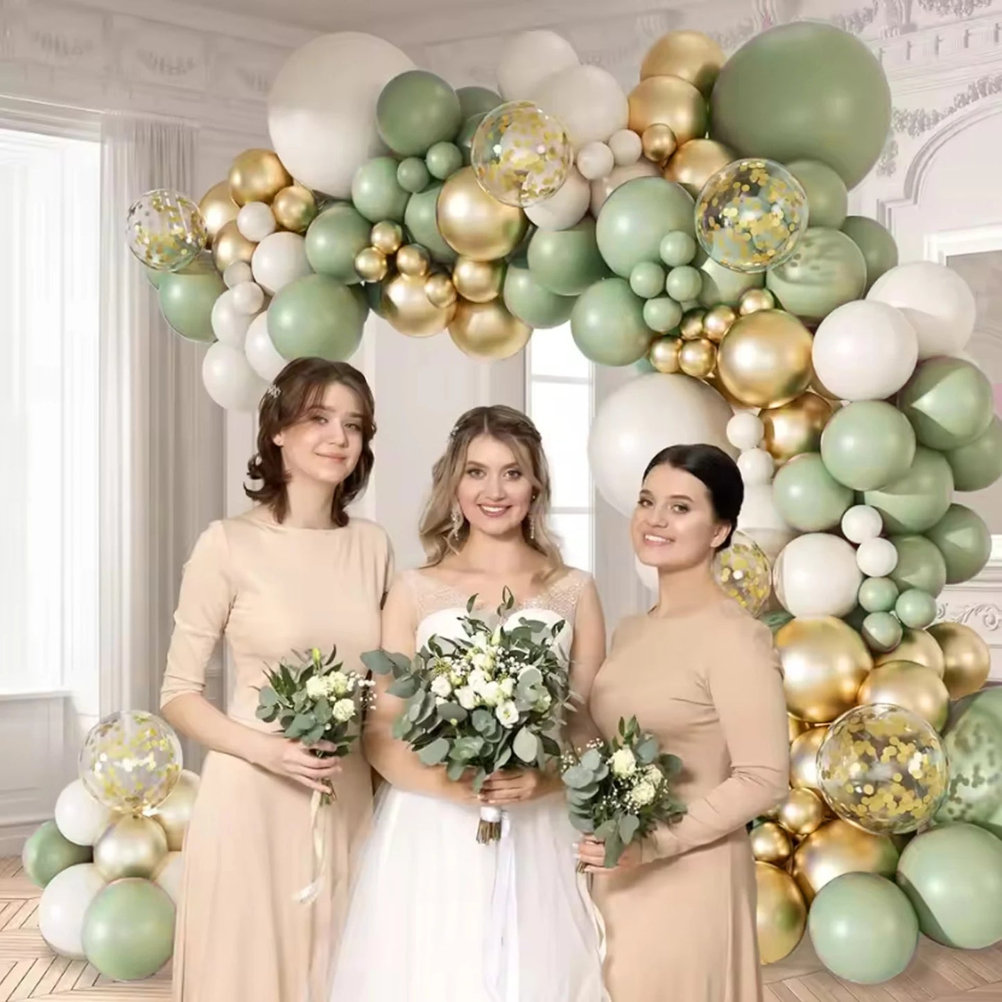 Three women posing in front of a large balloon arch decorated with sage green, white, and gold balloons, including clear balloons filled with gold confetti. The women are wearing matching beige dresses, and the bride holds a white bouquet.