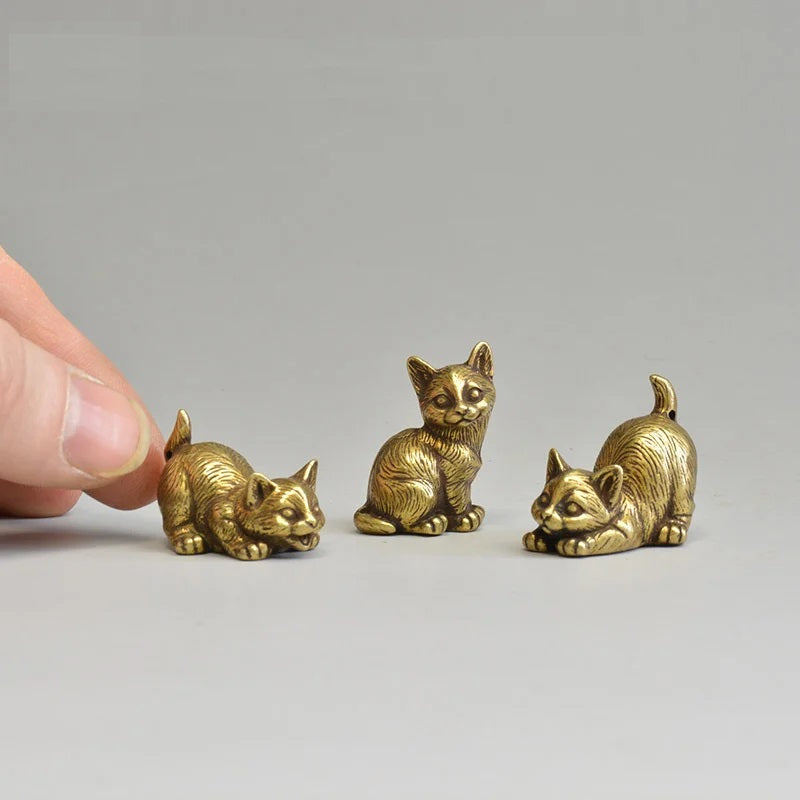 Three small brass cat figurines on a gray surface with a hand for scale.