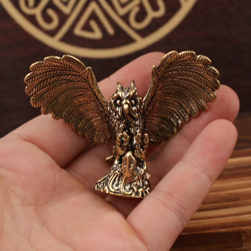 A small metallic owl figurine with spread wings, held in a person's hand. The owl has an antique gold/bronze finish with detailed feather and facial textures.