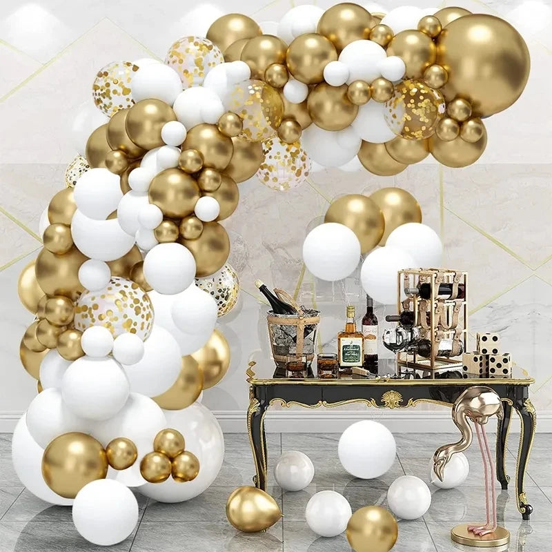 A luxury party decoration setup featuring a large balloon arch in metallic gold, white, and confetti-patterned balloons, paired with a matching gold and white bar cart holding bottles and serving accessories.